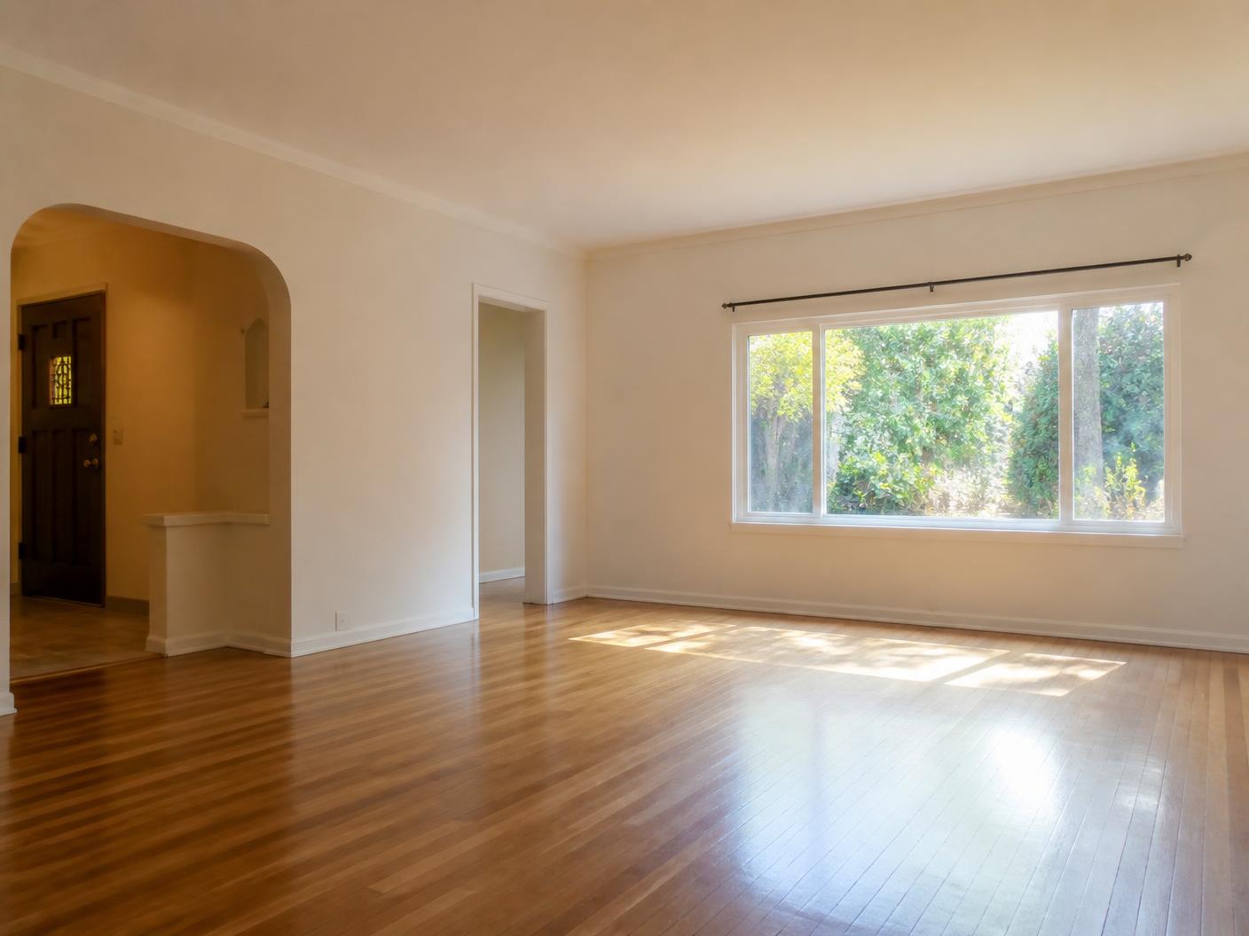 A bright, empty living room with wood floors and sunlight through a large window